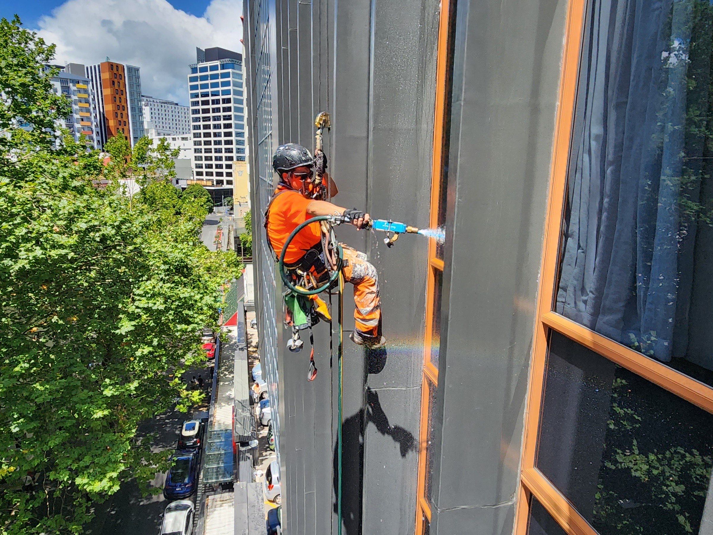 On Ropes technician washing a dark facade with a pressure lance, street trees below