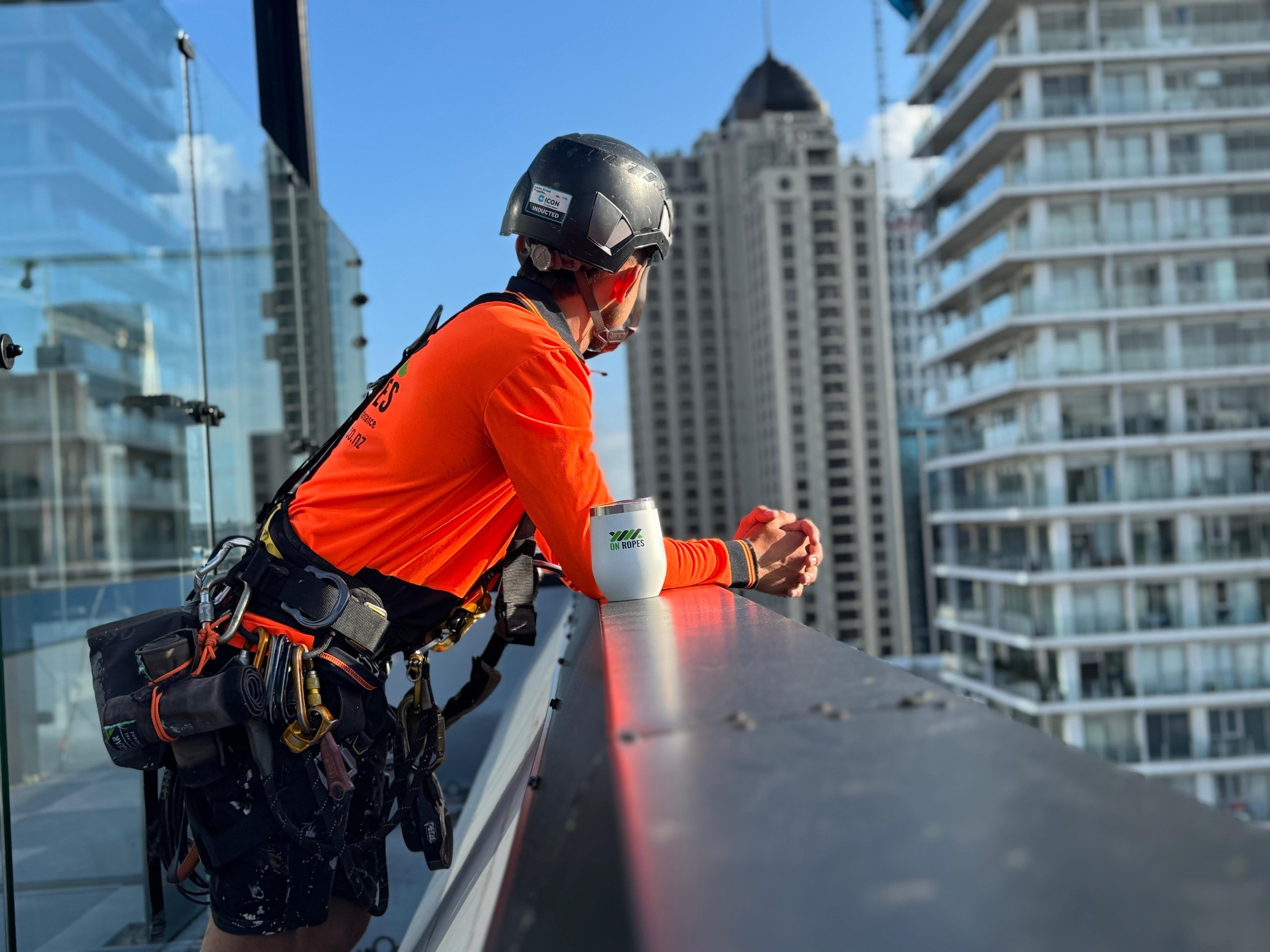 On Ropes technician resting at a rooftop parapet with a flask, Auckland apartment towers behind