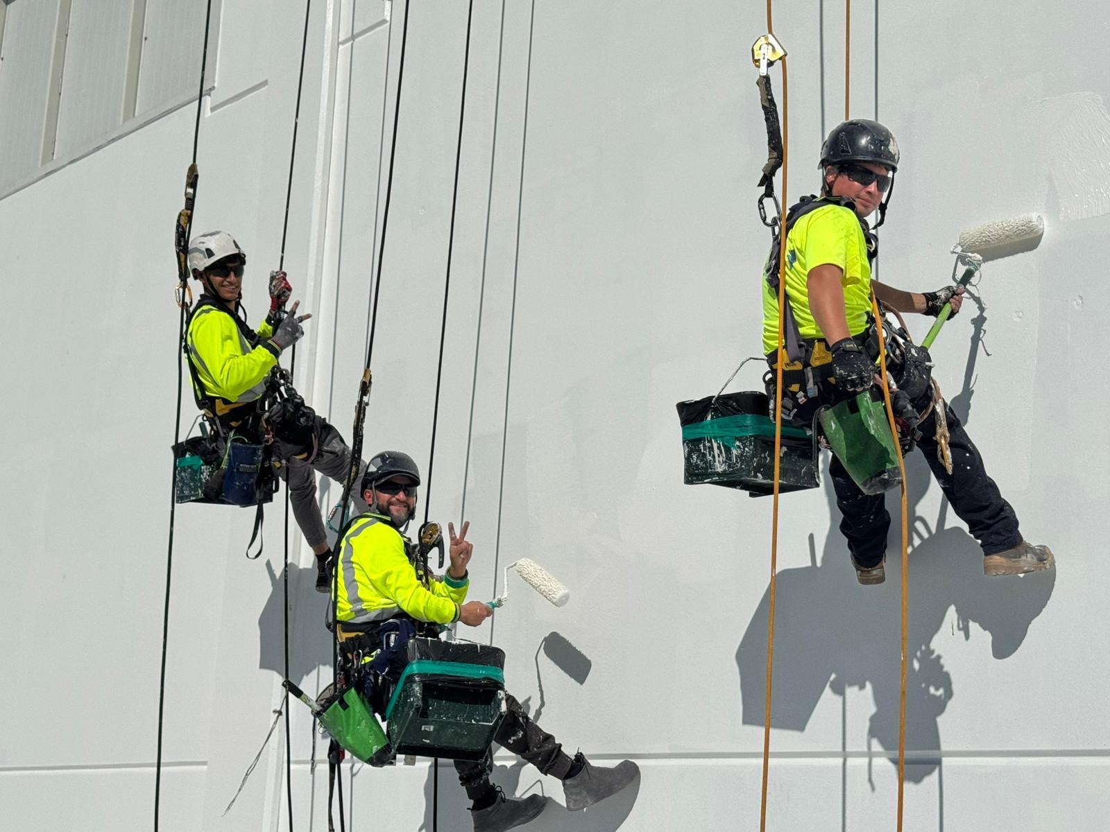 Three On Ropes technicians rolling a two-coat system across a white elevation from the rope