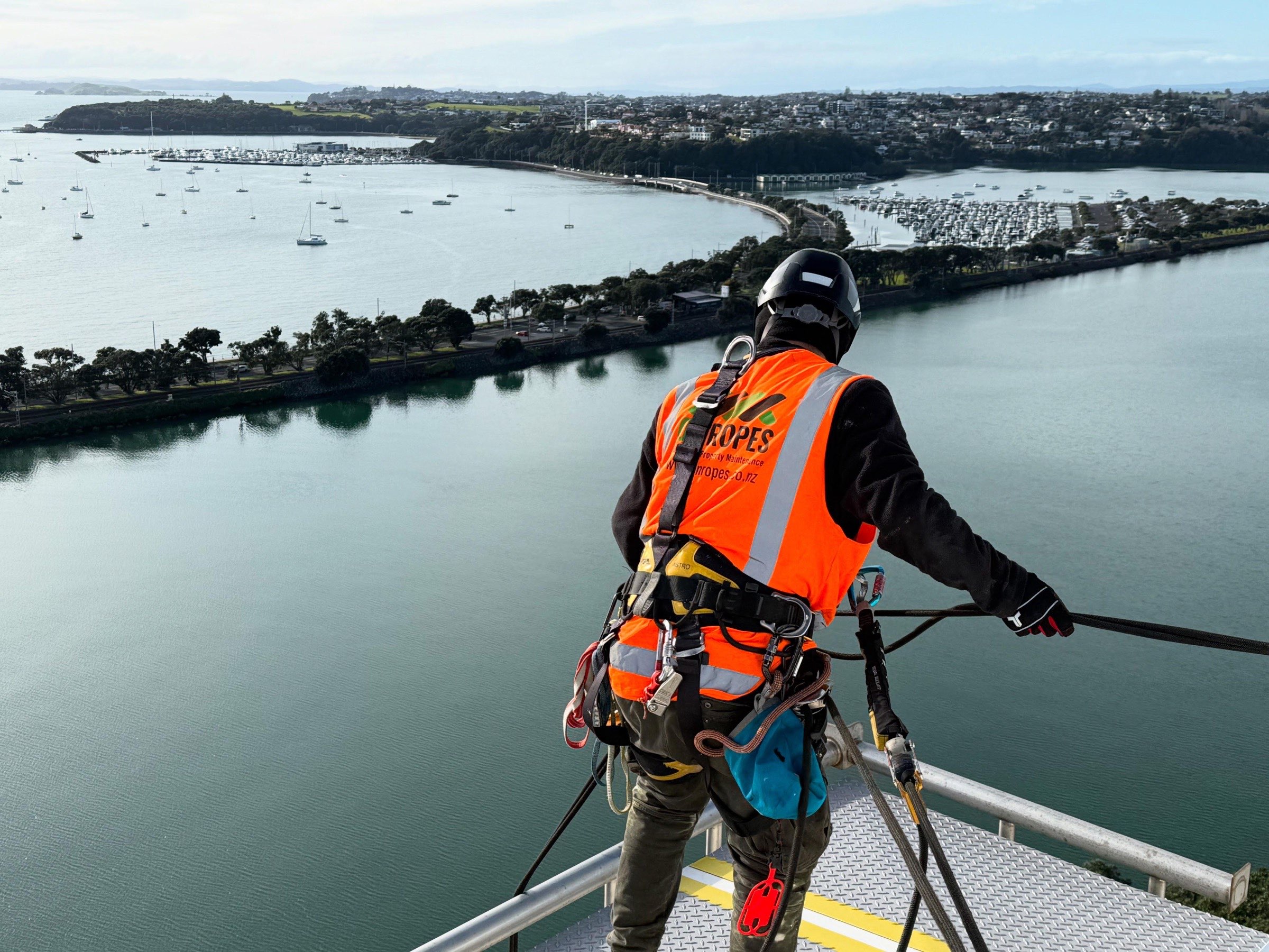On Ropes technician surveying from a rooftop edge above the Waitematā