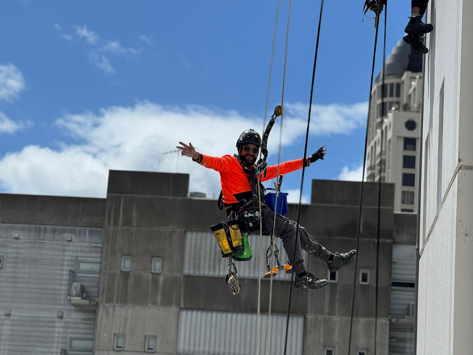 On Ropes technician hanging on twin ropes with both arms out, Auckland CBD behind
