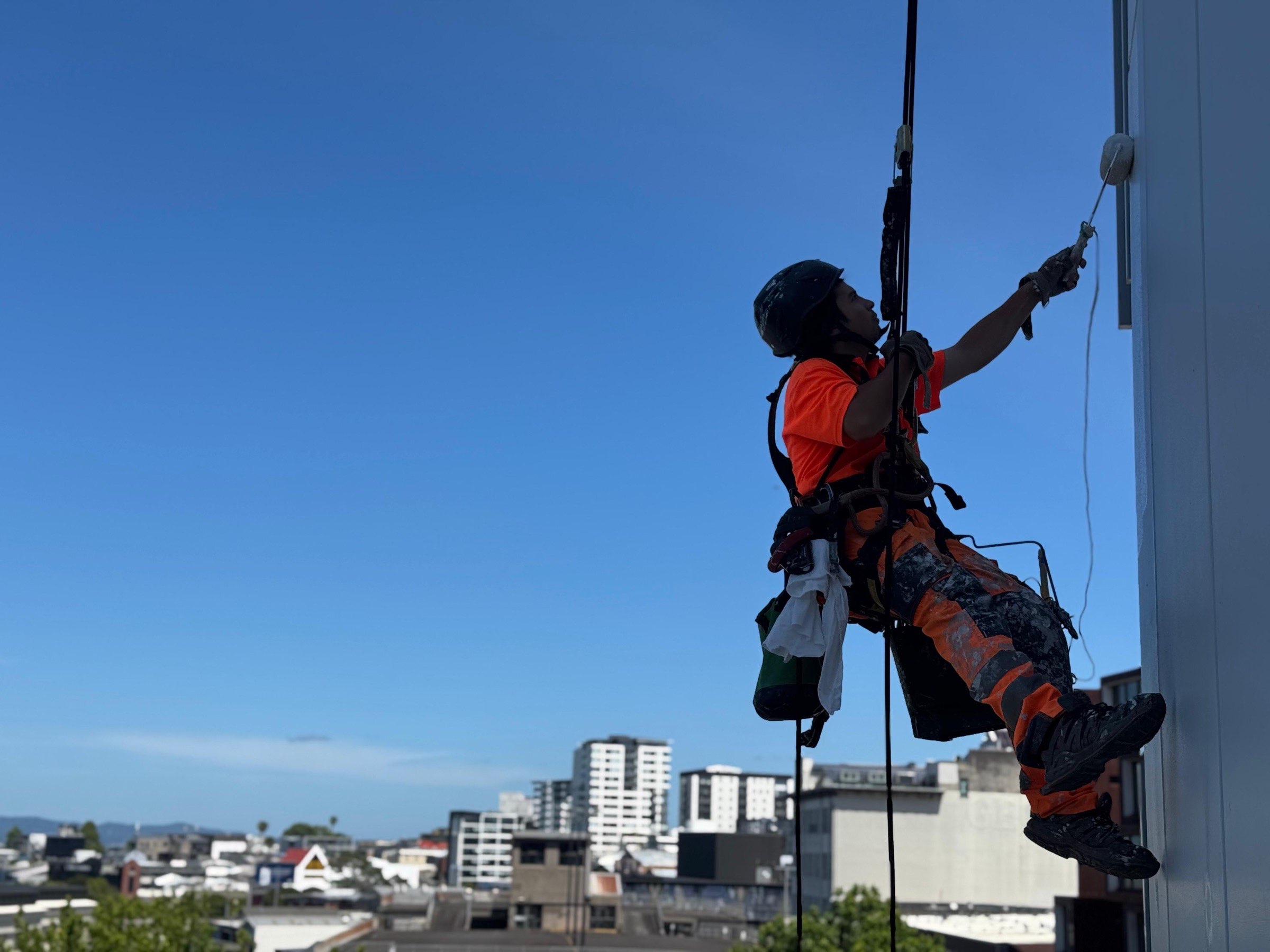 On Ropes technician rolling a coat onto an exterior wall from twin ropes, Auckland skyline behind